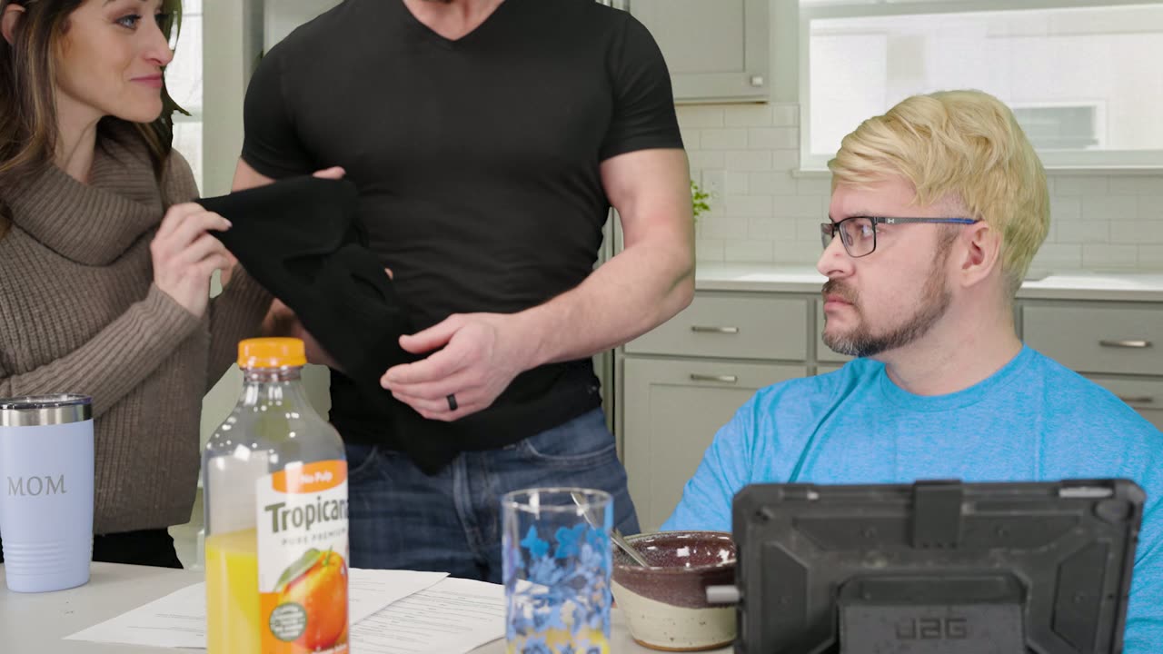 The man in the black t-shirt holding up a black cloth beside the woman while the man in the blond wig looks on
