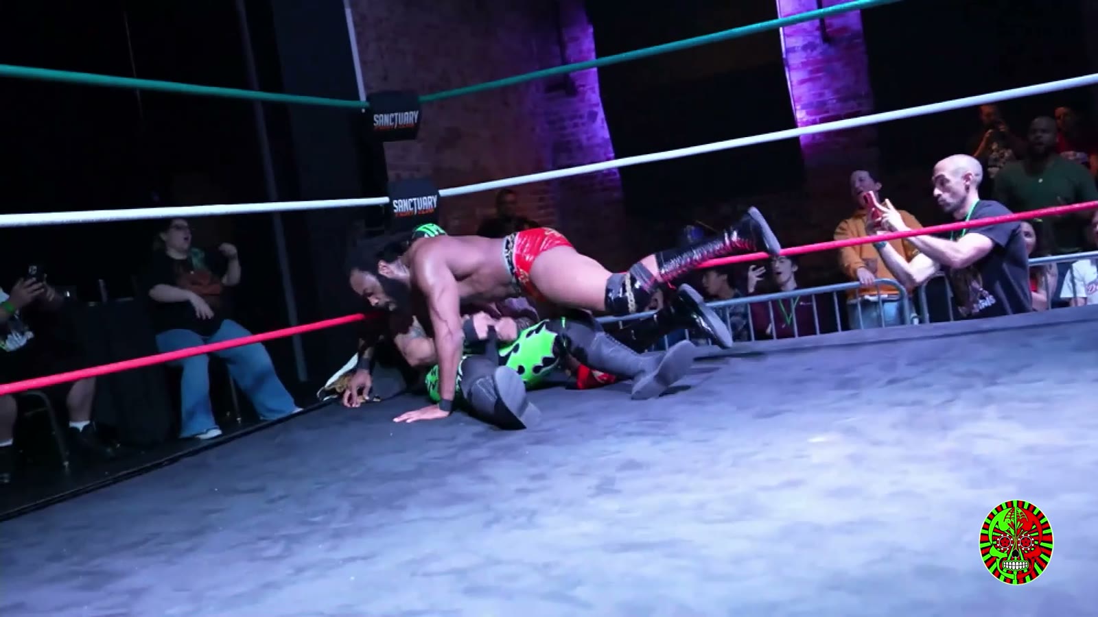 Low camera angle under the second rope at Lucha Boom: one wrestler on top of another on the mat, with fans filming behind