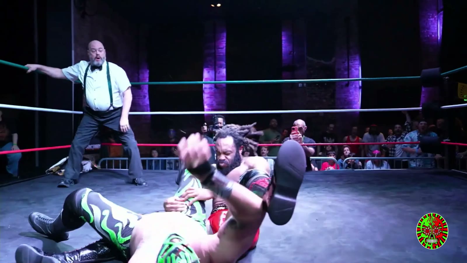 Low ringside shot at Lucha Boom of a wrestler straining in a hold on the mat, with the referee gesturing behind him